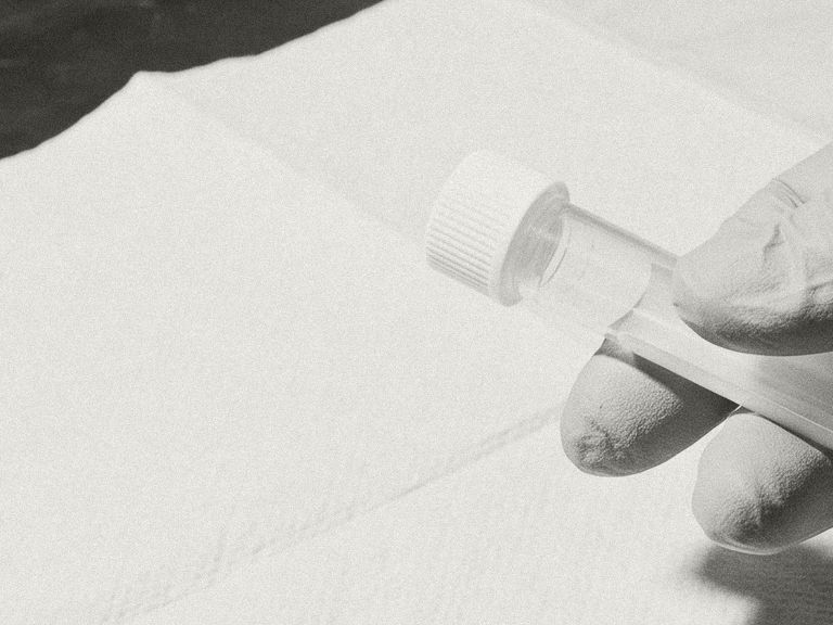 Black and white photograph of gloved fingers holding a capped sample tube over paper towel.